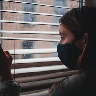 Woman in mask looks out window through blinds.