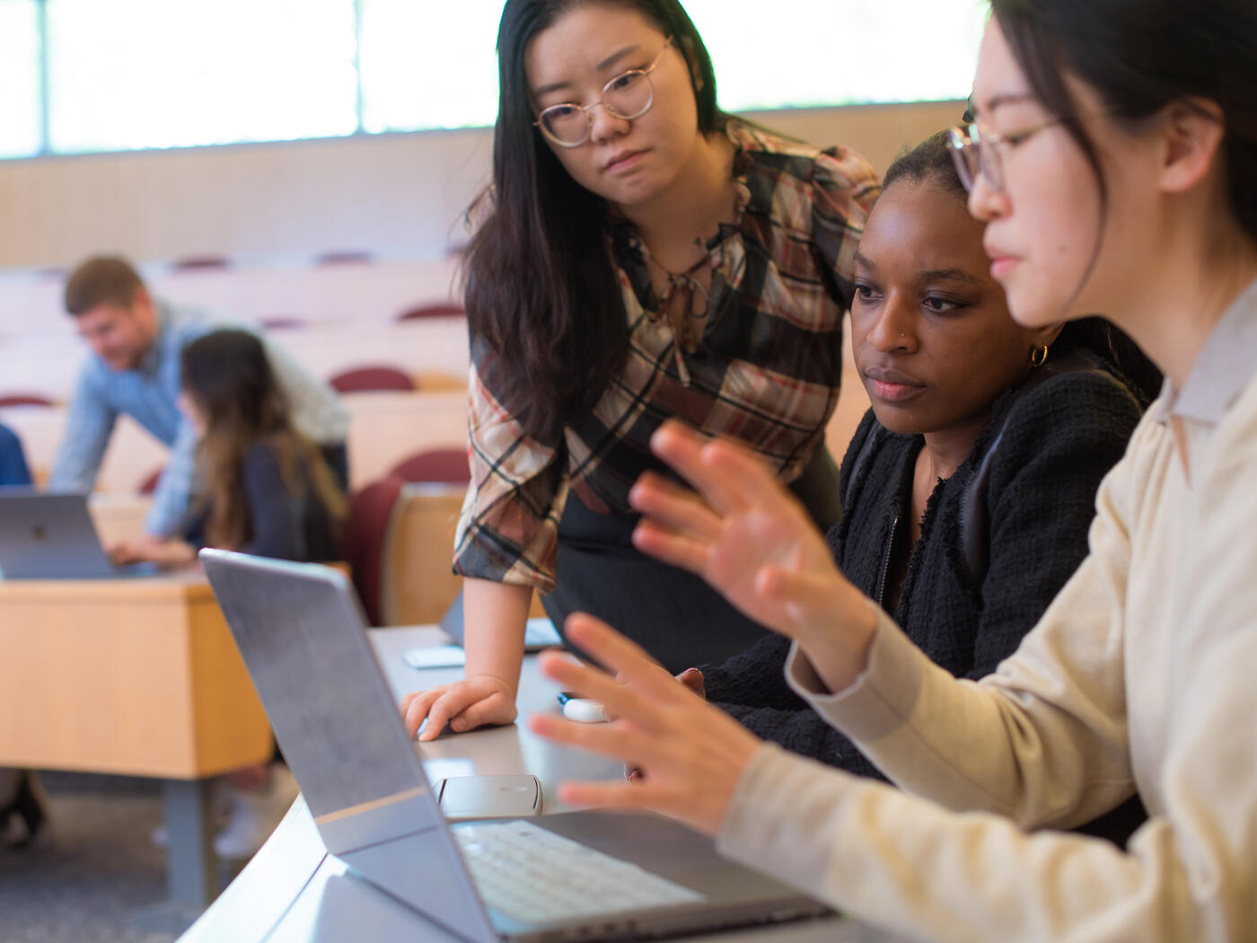 A group of three women looking at a laptop in a classroom and a group of three students in the background