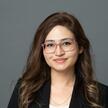 A headshot of Hibah, she in front of a dark grey background wearing pink clear frame glasses, a black blazer, and a white top. She has long brown hair.