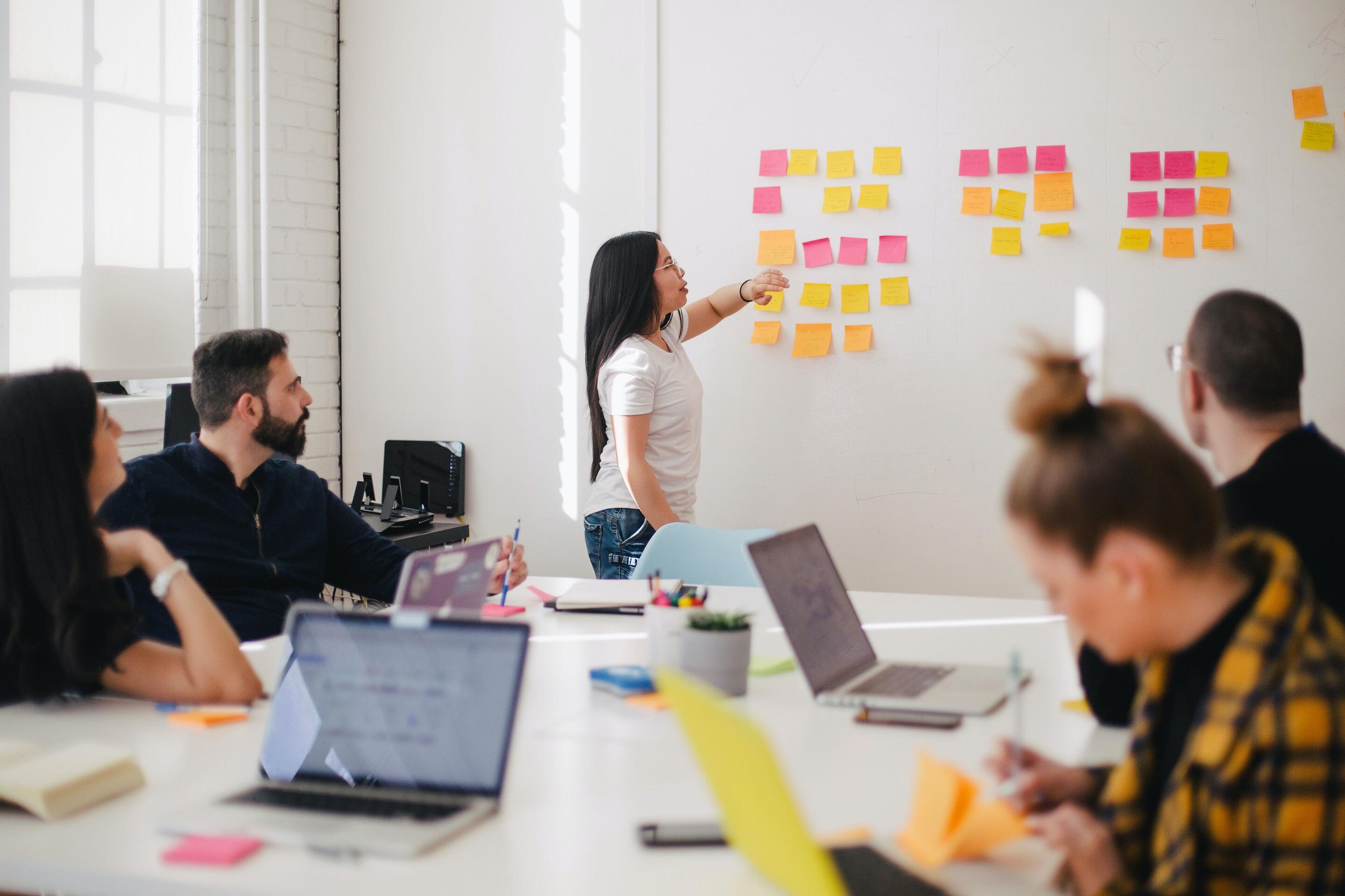 People having a meeting around a table, standing woman pointing to sticky notes on wall